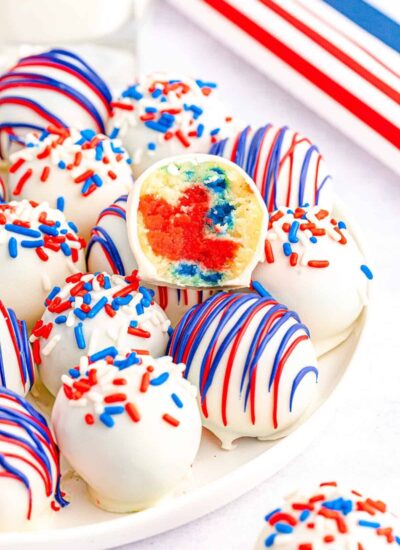 A plate of white chocolate-coated cake balls decorated with red, white, and blue sprinkles and drizzles. One cake ball is cut in half, revealing a colorful interior with distinct red and blue hues.