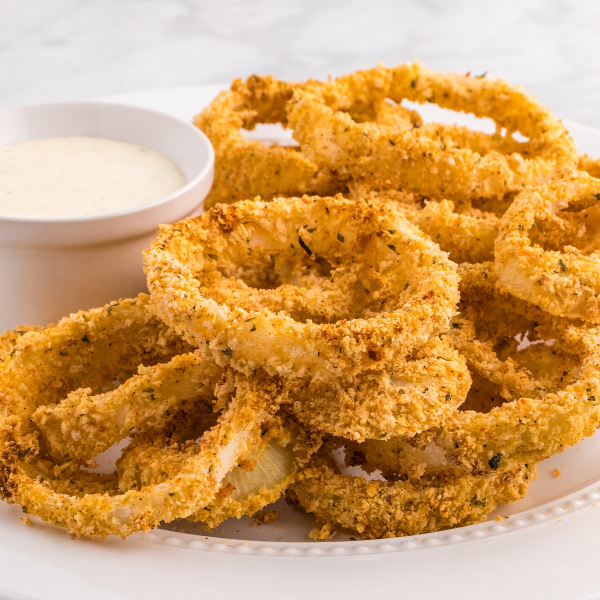 A plate of crispy, golden-brown Air Fryer onion rings beside a small bowl of dipping sauce.