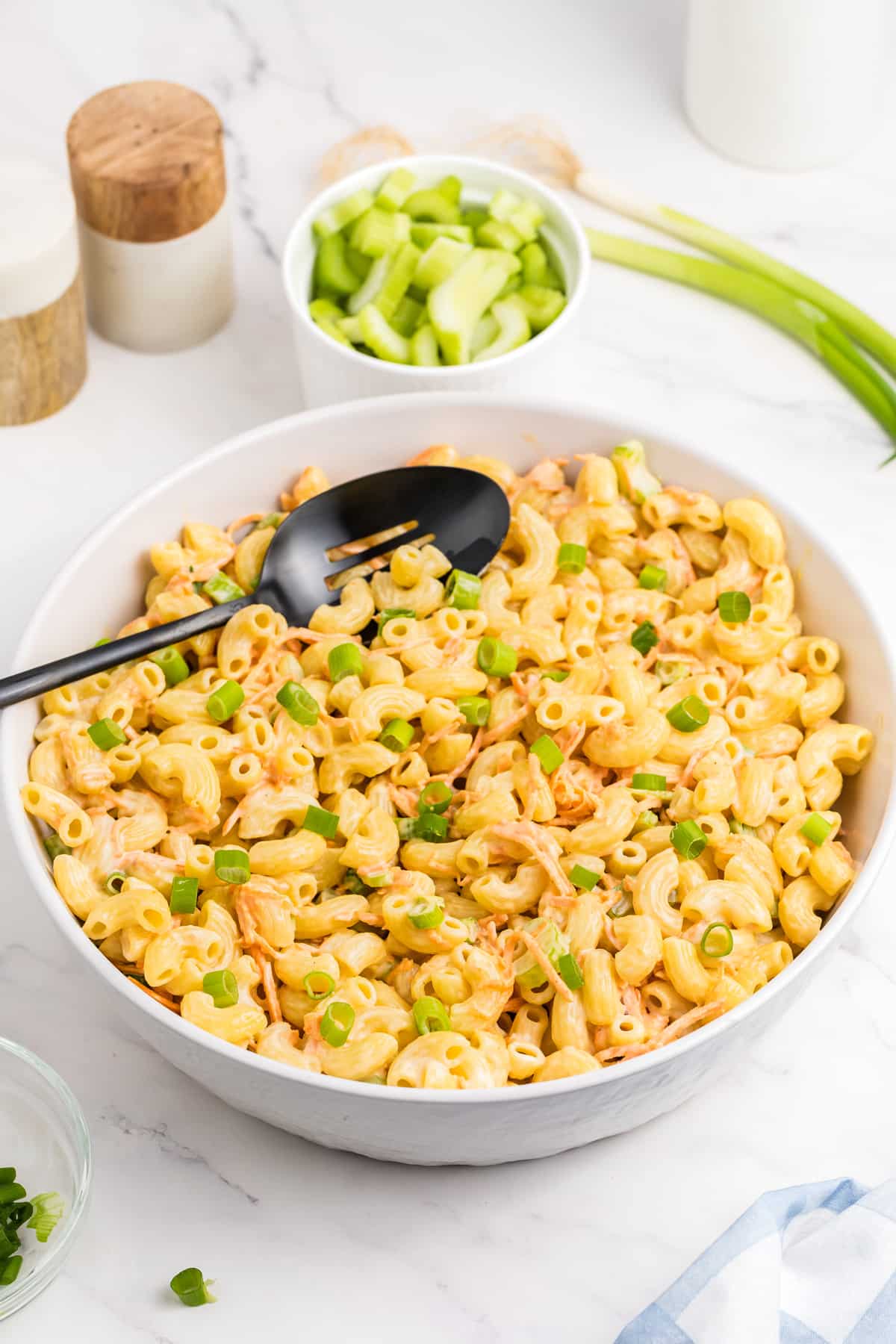 A bowl of Hawaiian Macaroni Salad garnished with chopped green onions, placed on a marble surface. There is a black serving spoon in the bowl and a small bowl of chopped celery nearby.