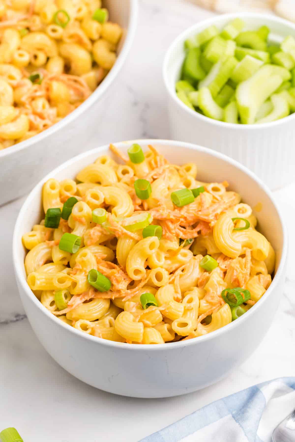 A bowl of Hawaiian Macaroni Salad with shredded carrot and chopped green onions, placed on a marble surface. A small bowl of sliced celery is in the background.