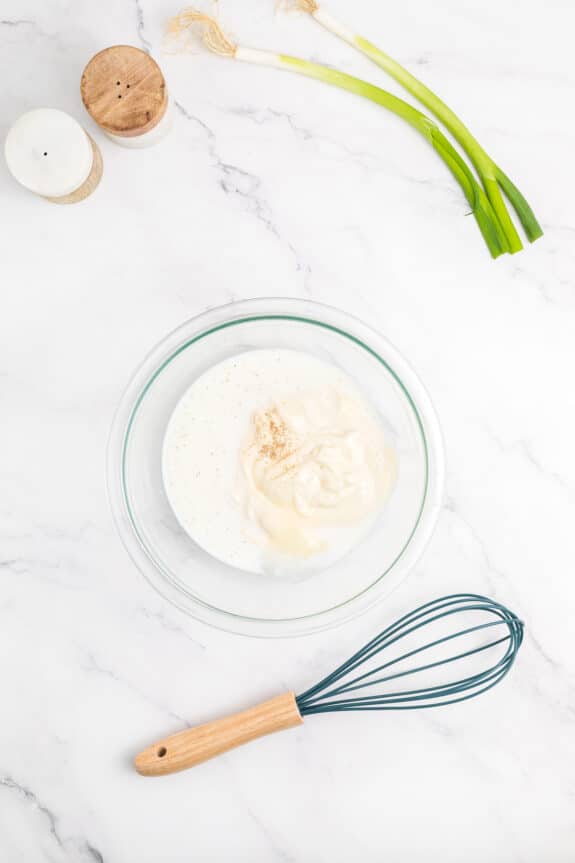 Hawaiian Macaroni Salad recipe process is to use a glass bowl containing mayonnaise and seasoning on a marble countertop. Nearby are spring onions, a whisk, and two salt shakers.