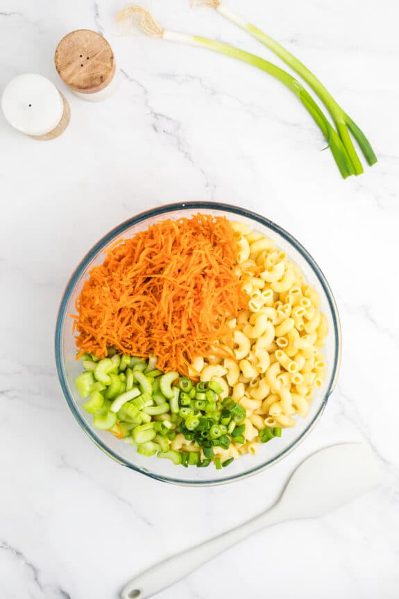 Hawaiian Macaroni Salad ingredients in a glass bowl with shredded carrots, macaroni, and chopped celery. A white spatula is beside it on a marble surface, with green onions and salt and pepper shakers in the background.