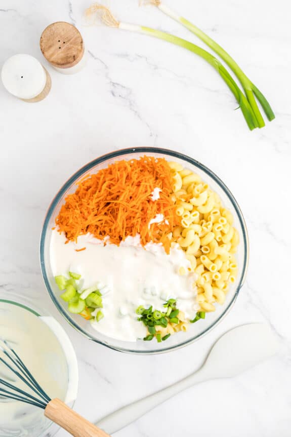 Mixing the ingredients for Hawaiian Macaroni Salad in a bowl. A blend of macaroni,shredded carrots, chopped green onions, and creamy dressing on a marble surface. Whisk, spatula, salt, pepper shakers, and green onions nearby.