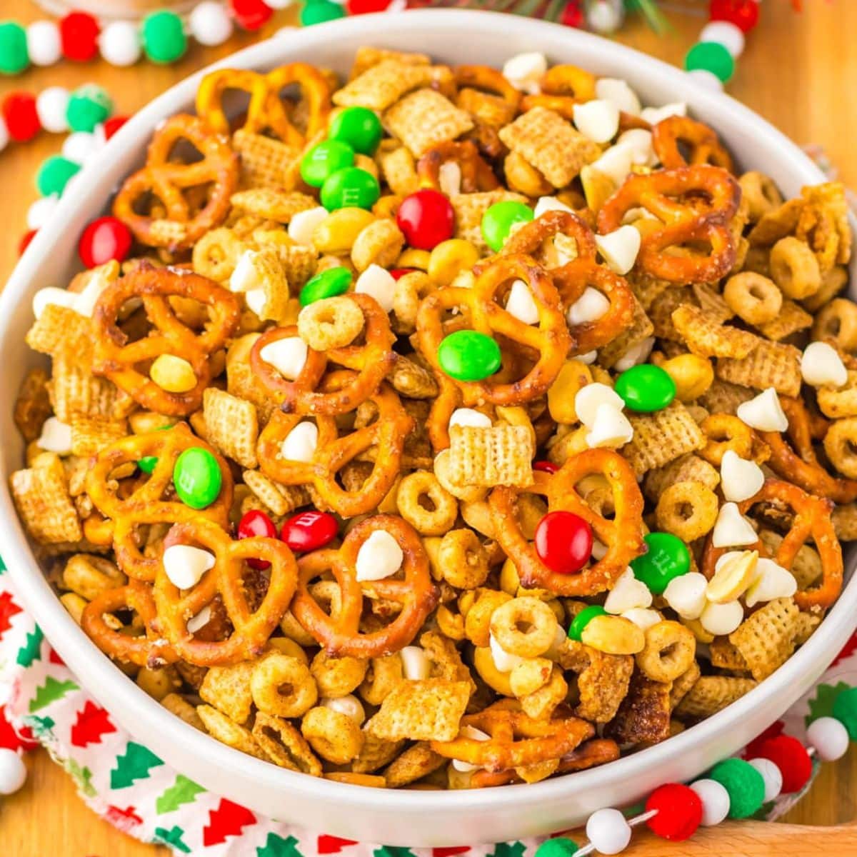 A bowl filled with a snack mix of pretzels, cereal, candy-coated chocolates, white chocolate chips, and peanuts. A festive, colorful beaded decoration surrounds the bowl.