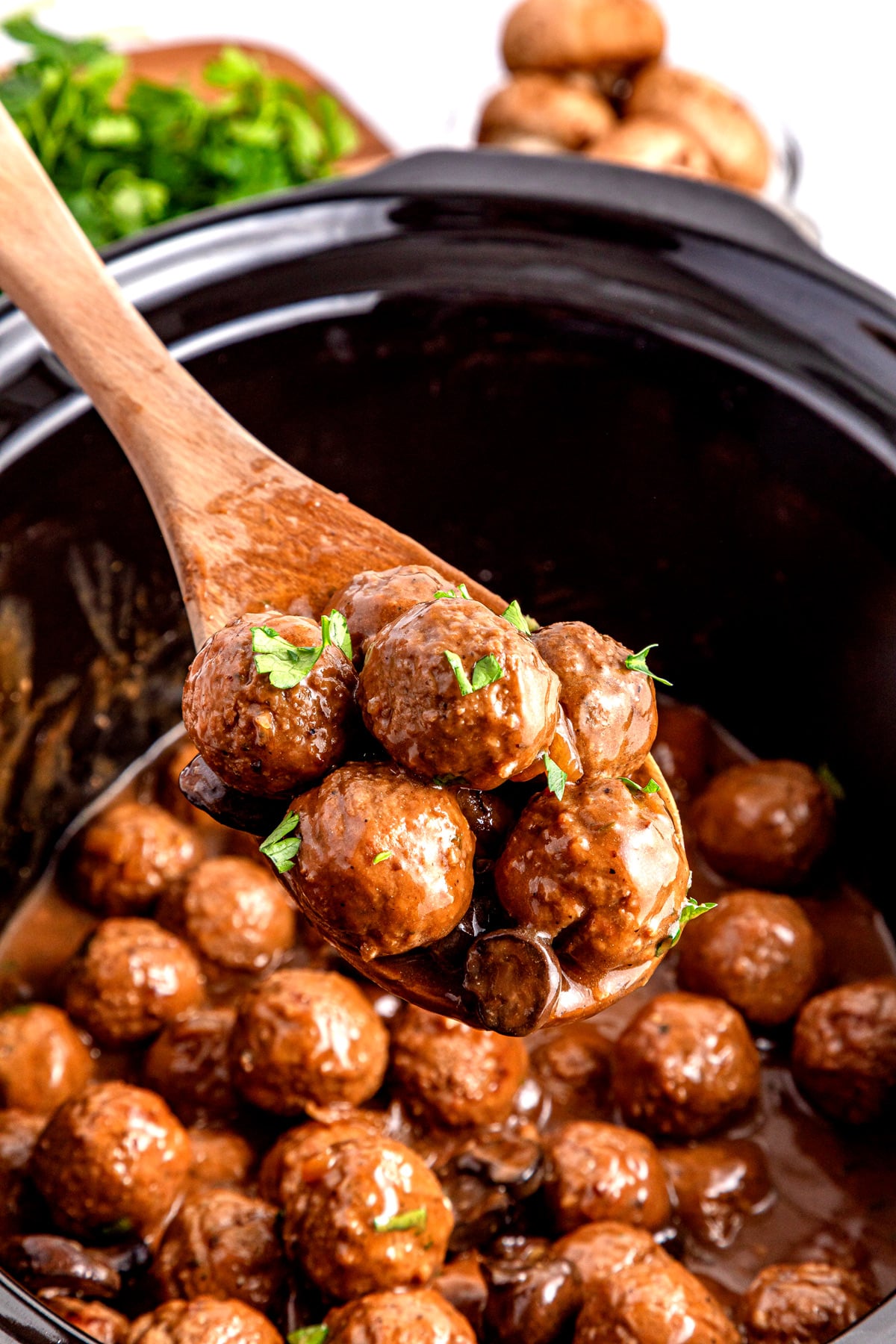 A wooden spoon holds Salisbury Steak Meatballs covered in sauce, with chopped herbs, over a slow cooker filled with more meatballs.