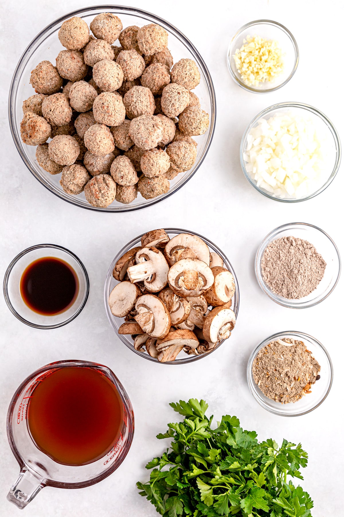 Ingredients for a dish, including bowls of meatballs, sliced mushrooms, chopped onions, minced garlic, beef broth, worcestershire sauce, flour, and fresh parsley, brown gravy, kosher salt, chopped onion arranged on a marble surface.