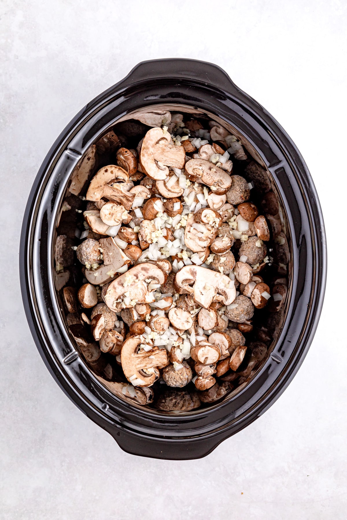 Top view of a slow cooker filled with sliced mushrooms, chopped onions, and small potatoes for Salisbury Steak Meatballs recipe.
