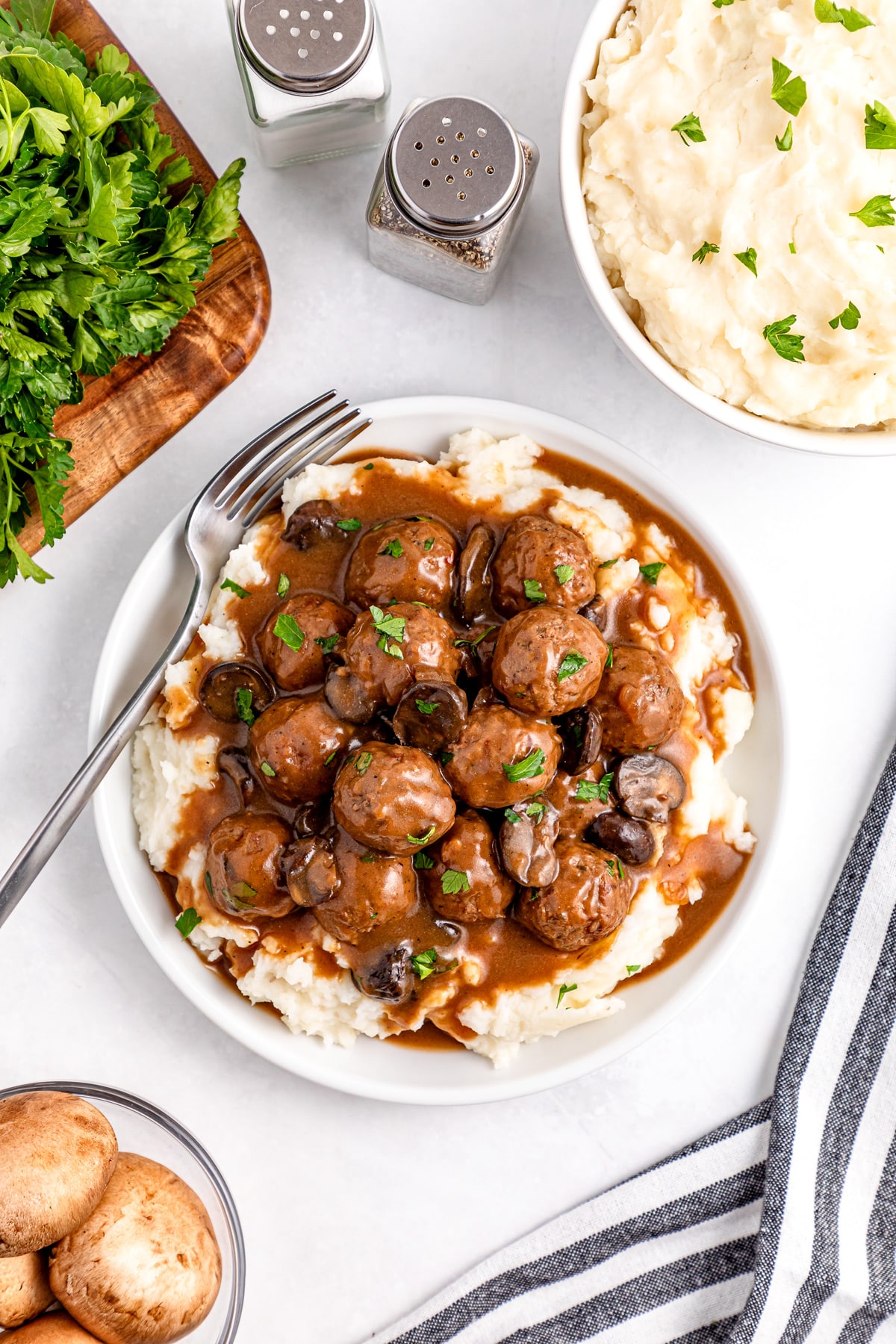 Salisbury Steak Meatballs in a plate with gravy on mashed potatoes, garnished with parsley. Nearby are mashed potatoes, fresh parsley, salt and pepper shakers, mushrooms, and a striped cloth.