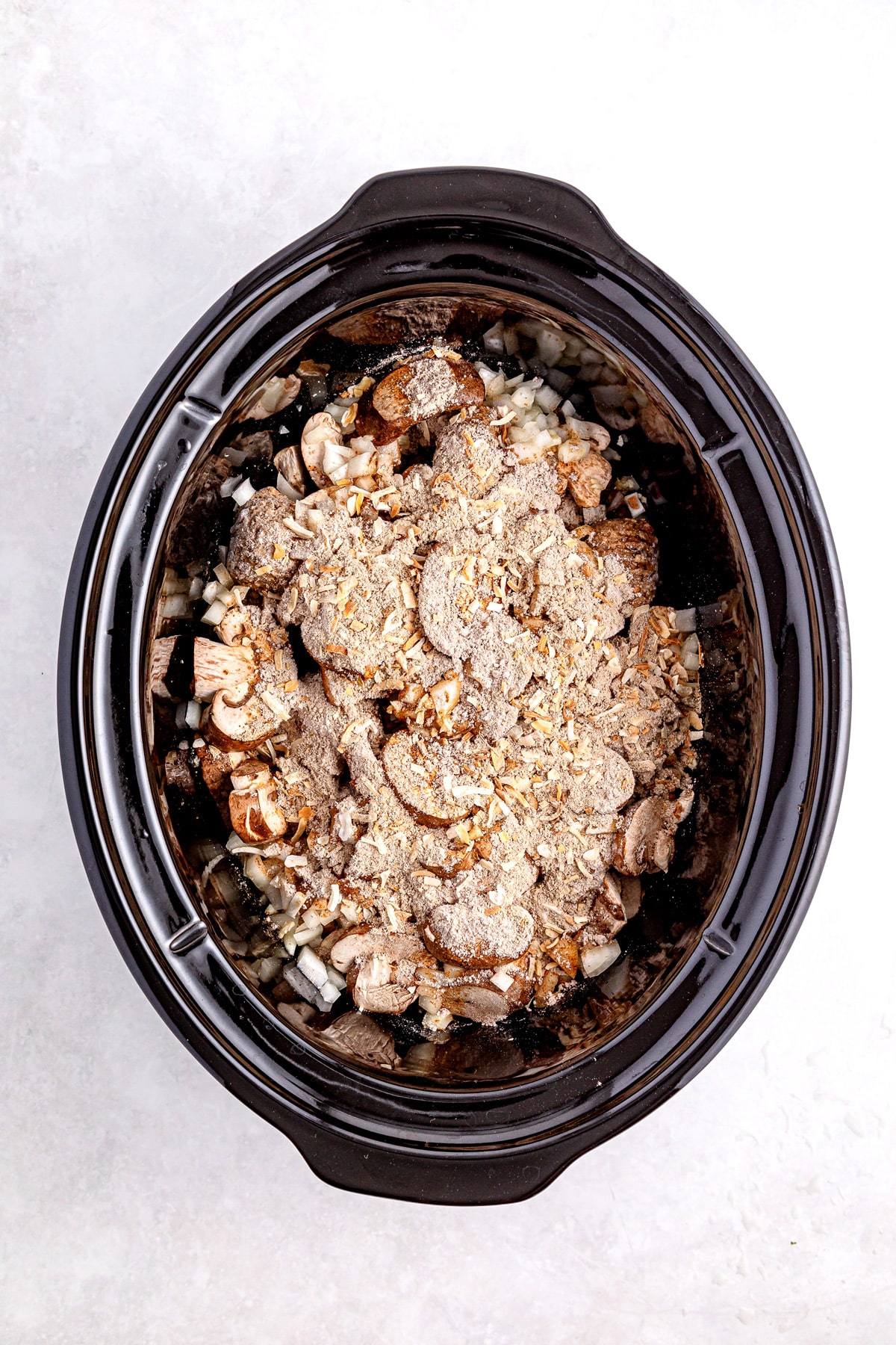 Filling the slow cooker with sliced mushrooms, diced onions, and seasoning on top of raw ingredients as a step in making Salisbury Steak Meatballs
