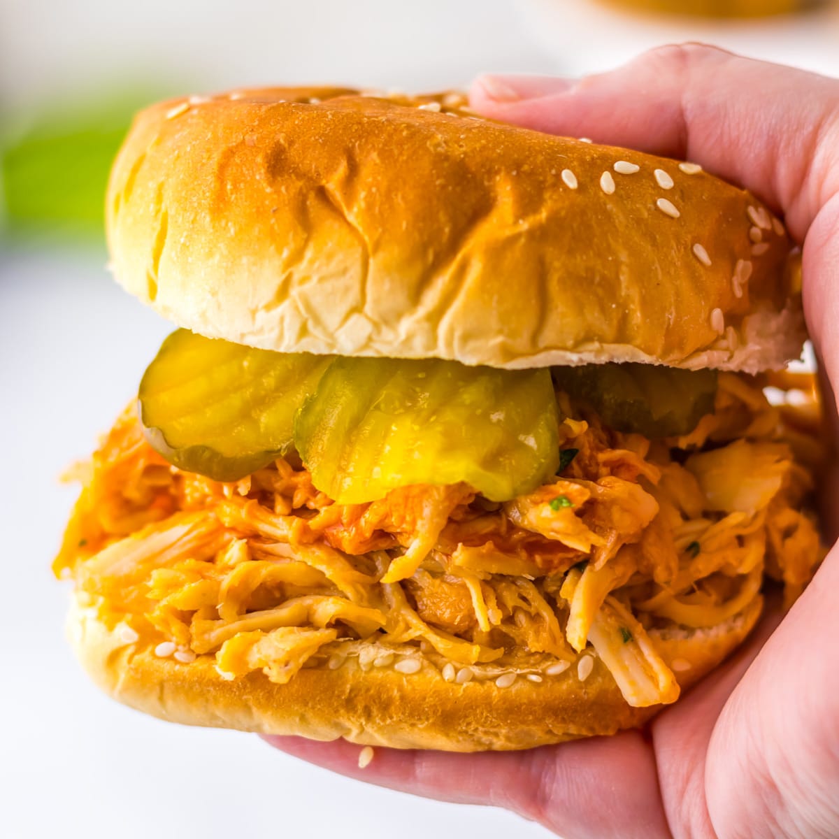 A hand holding a Slow Cooker Nashville Hot Chicken sandwich on a sesame seed bun with pickles.