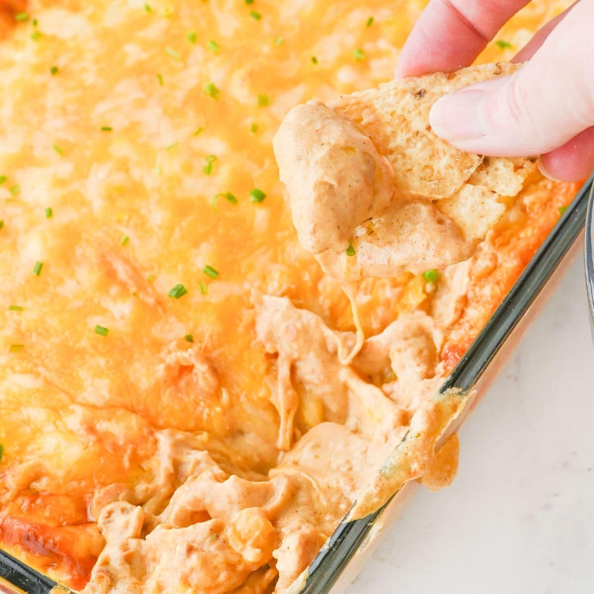 Hand dipping a tortilla chip into a cheesy baked dip in a glass dish.