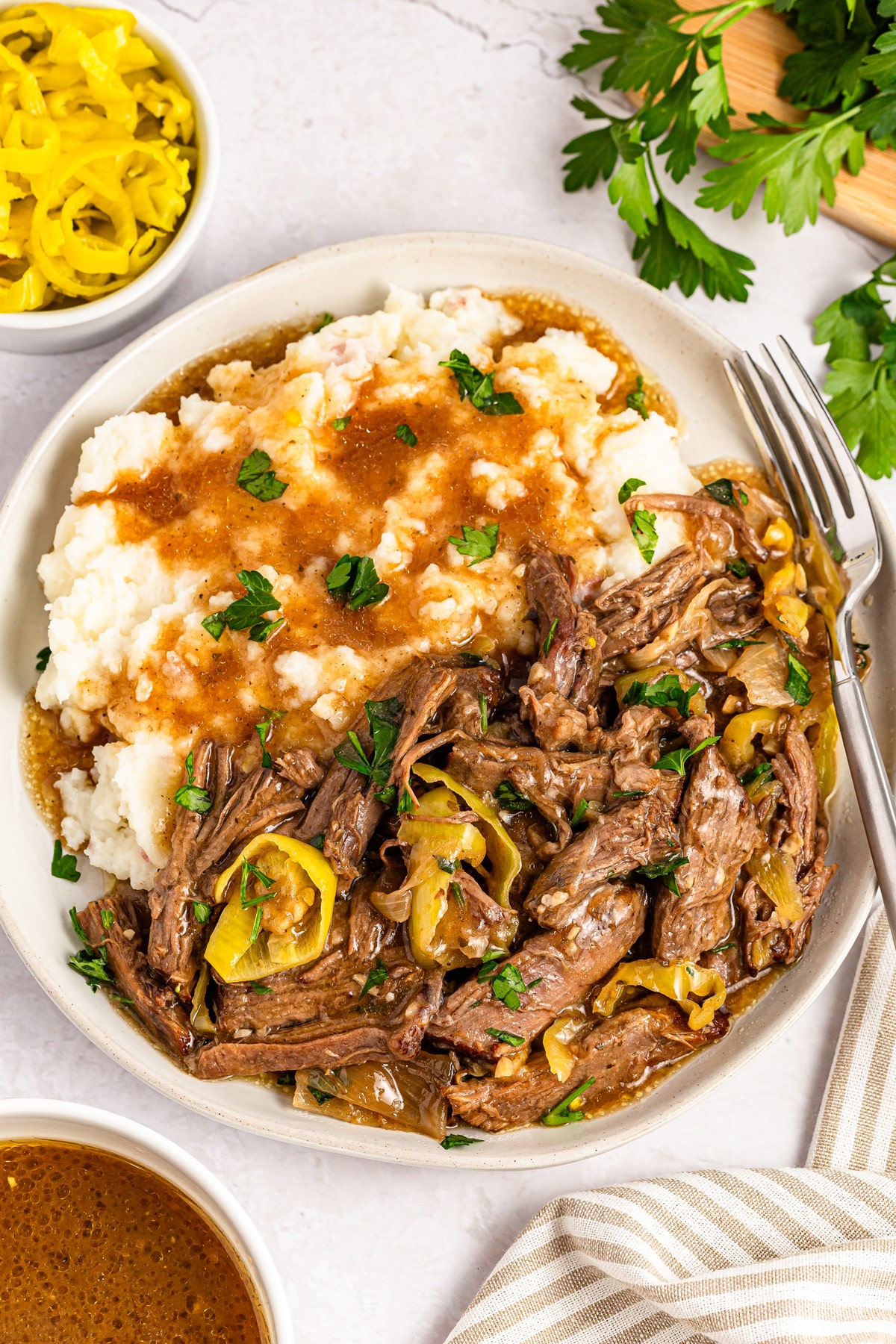 Plate of shredded beef with peppers and gravy next to mashed potatoes, garnished with parsley. Fork on the side. Bowls of sauce and pepper slices nearby, with a sprig of parsley above.