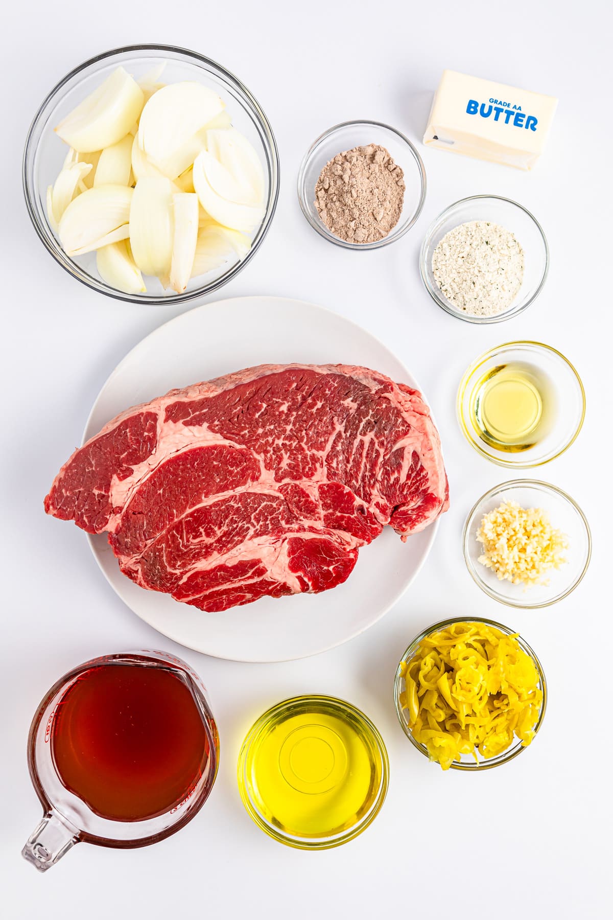 A spread of ingredients including raw beef, sliced onions, butter, sliced pepperoncini, broth, oil, garlic, seasonings, and a dry mix, arranged on a white surface.
