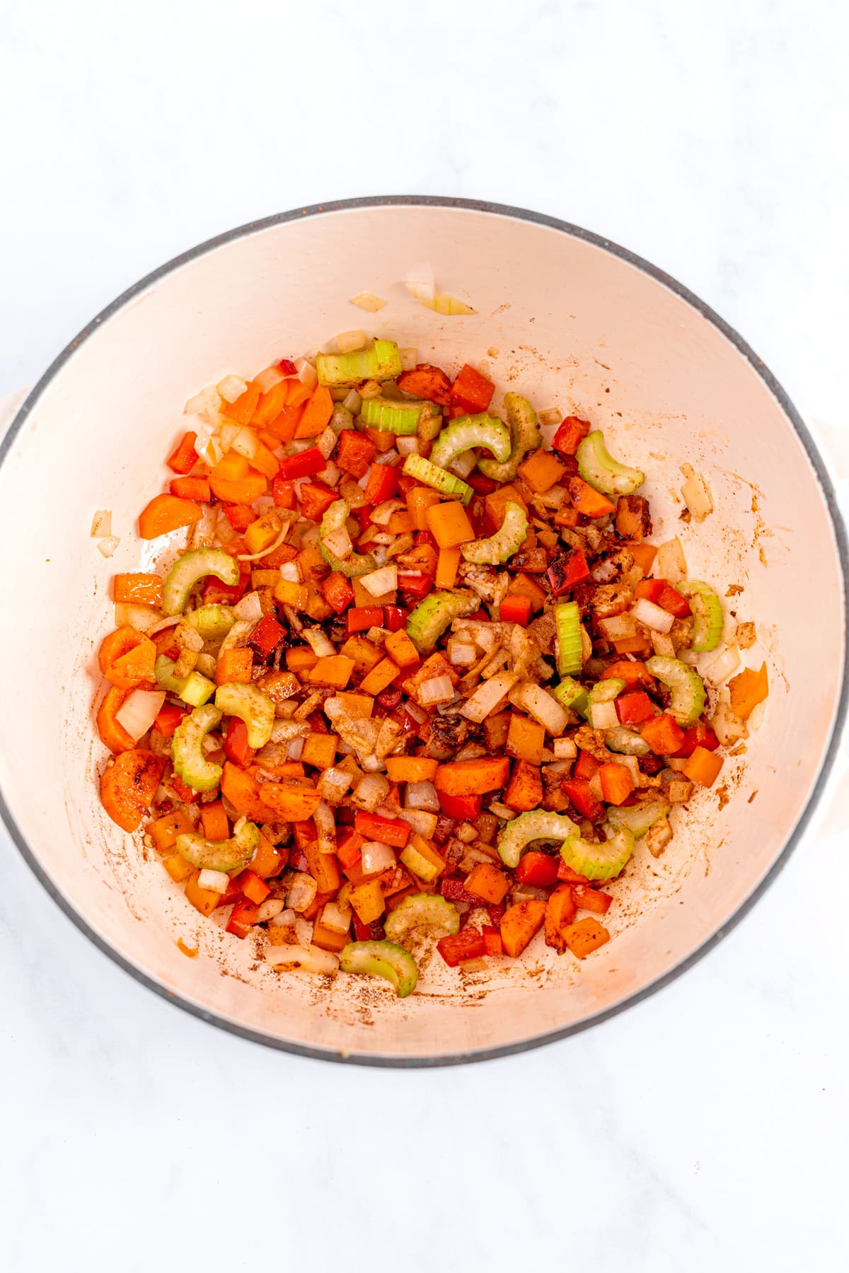 Chopped vegetables including onions, carrots, celery, and red peppers being sautéed in a white pot.