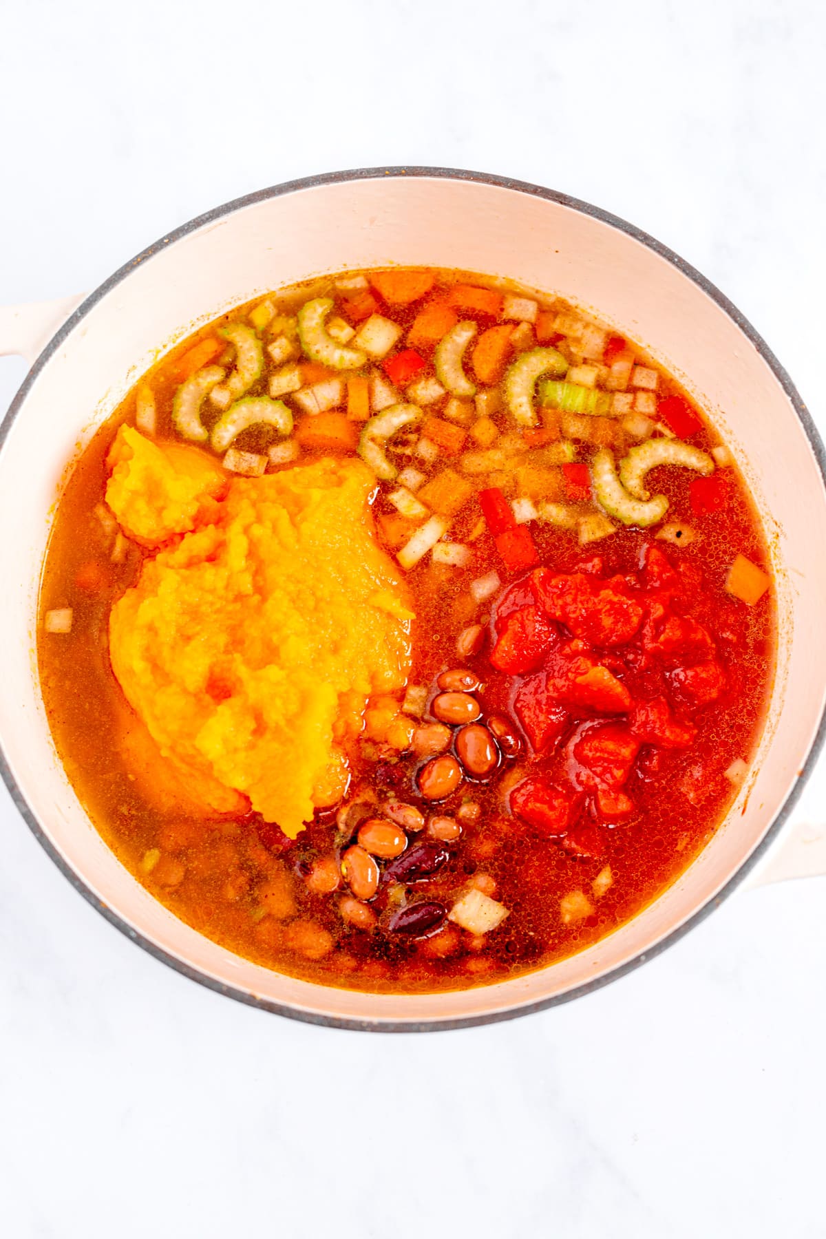 A pot filled with ingredients including tomato chunks, beans, celery slices, diced onions, and a scoop of mashed pumpkin, all in a liquid broth.