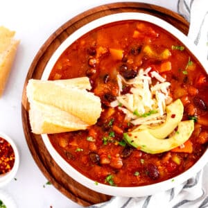 A bowl of Pumpkin Chili with black beans, topped with shredded cheese, avocado slices, and herbs. Two pieces of bread are beside it on a wooden plate.