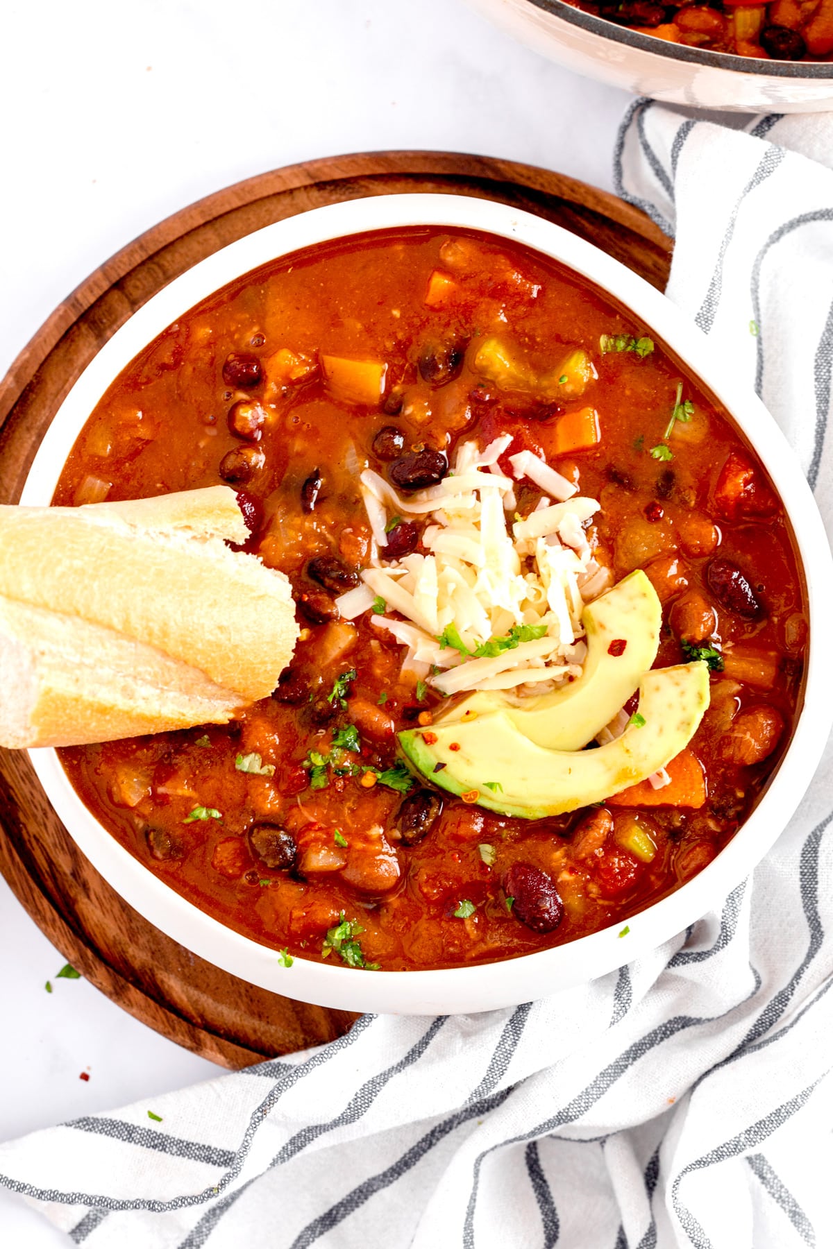 A bowl of Pumpkin chili topped with avocado slices, shredded cheese, and herbs, served with a piece of bread on the side, on a round wooden serving board.