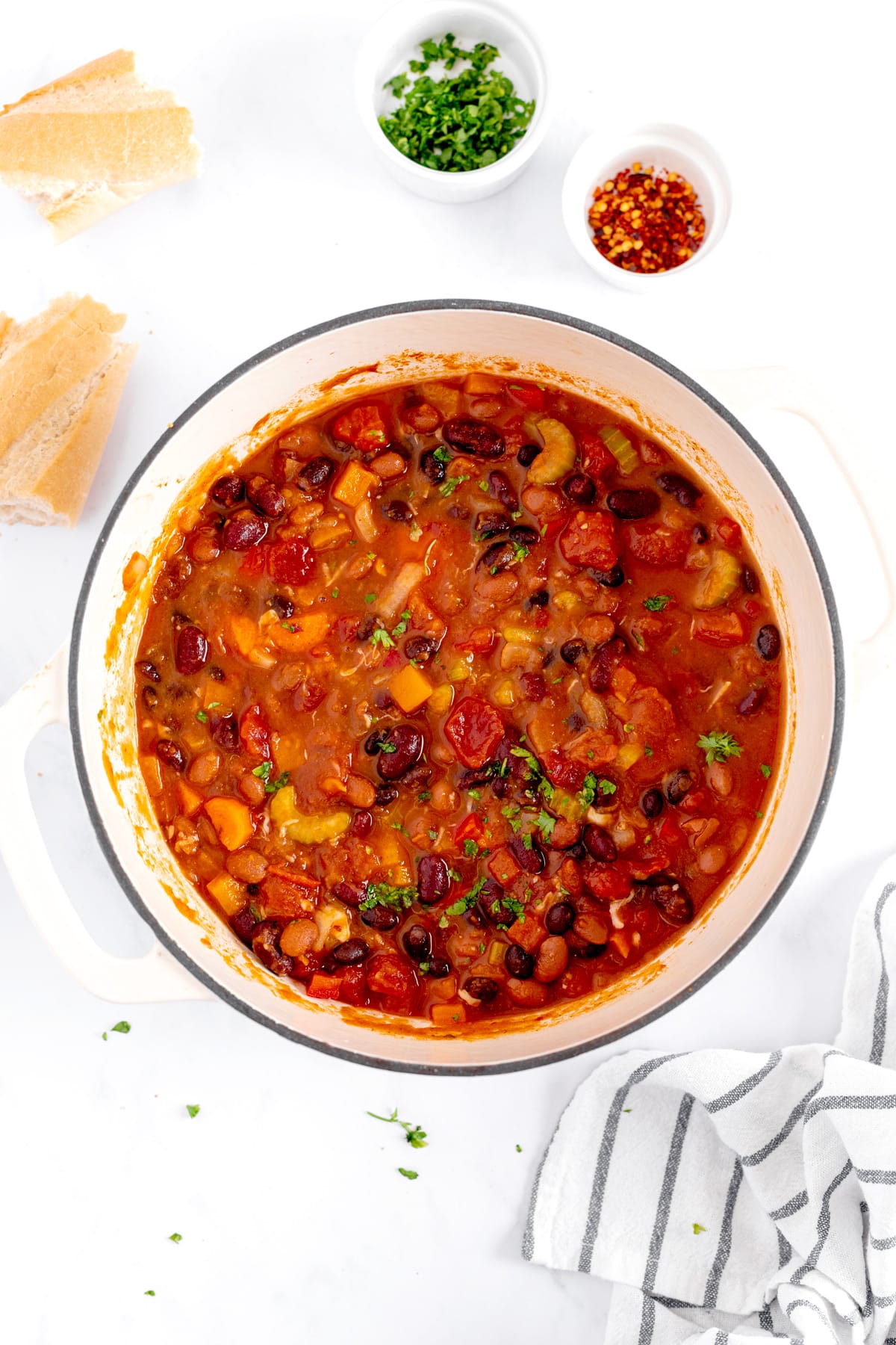 A pot of Pumpkin Chili with beans and tomatoes is on a stovetop. Nearby are bread pieces, a striped cloth, and small bowls of chopped herbs and chili flakes.