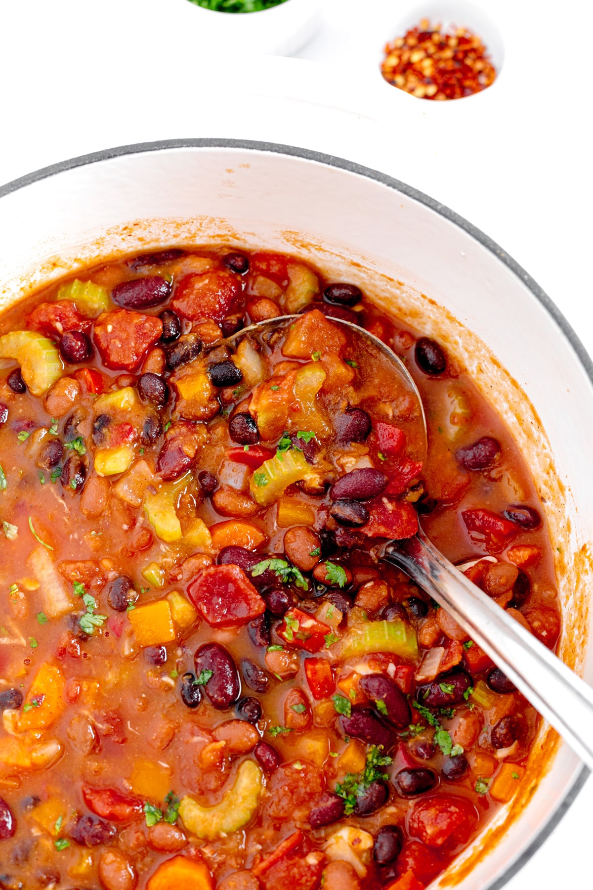 A pot of vegetable chili with beans, tomatoes, celery, and peppers, stirred by a ladle. Small bowls of red pepper flakes and chopped herbs are in the background.