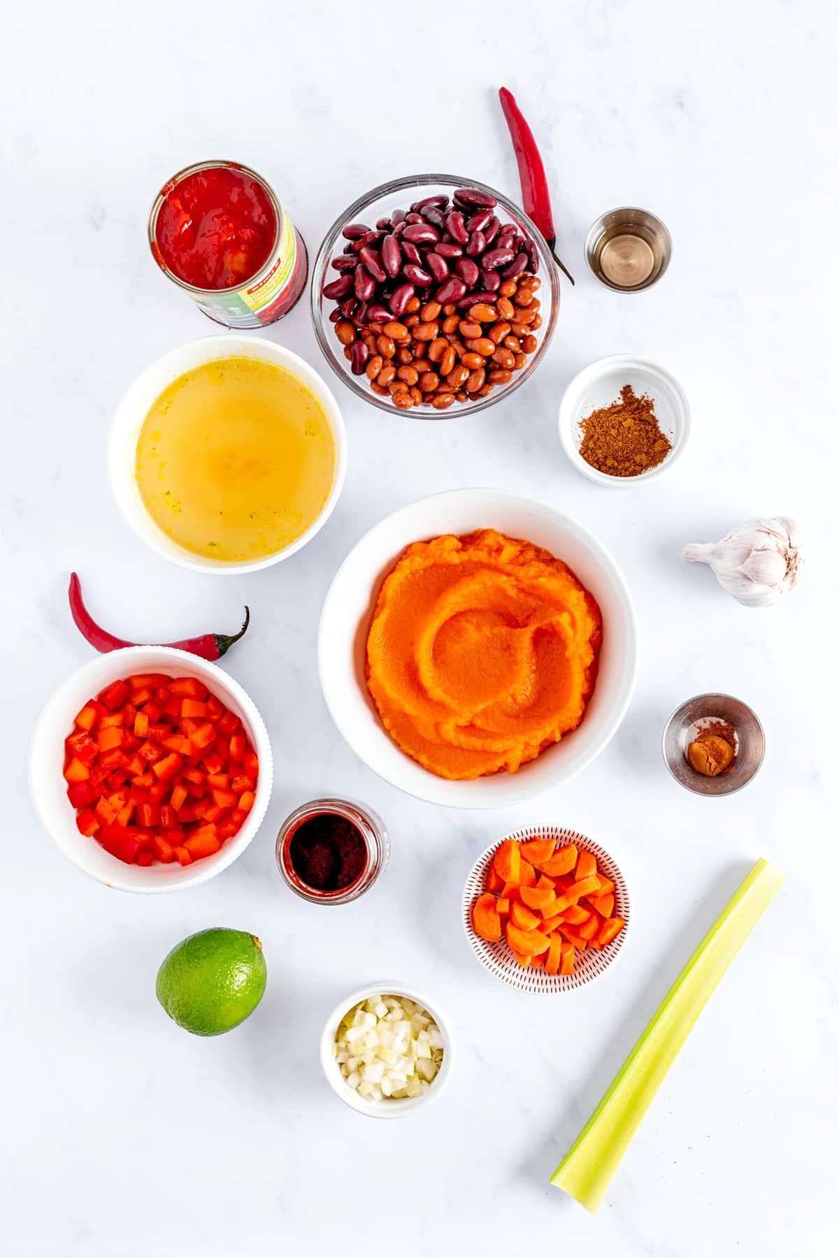 These are the ingredients for Pumpkin Chili, laid out on a white marbled counter: 1 cup of diced yellow onion, 2 tablespoons of neutral cooking oil, 1 finely diced hot red pepper (such as serrano, chipotle, habanero, or jalapeño, with seeds removed), and 2 teaspoons of minced fresh garlic. Season with salt, black pepper, ½ teaspoon of ground cinnamon, 1 teaspoon of chili powder, and 1 teaspoon of cumin. You'll also need 2 cups of vegetable broth (or any meat/poultry broth), 2 cups of canned chopped tomatoes (fire-roasted recommended), and 1 (15 oz) can of pumpkin puree (or homemade roasted pumpkin). Add 3 (15 oz) cans of beans (kidney, pinto, or your preferred variety), 1 large sliced carrot, 1-2 stalks of sliced celery, and 1 cup of diced red bell pepper. Serve with a splash of fresh lime juice.