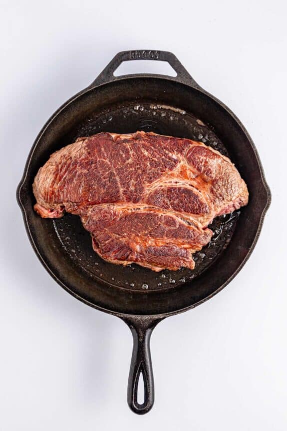 A raw steak with marbled fat is placed in a black cast iron skillet on a white background.