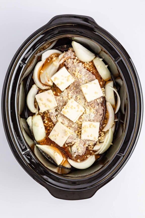 A slow cooker contains a raw beef roast topped with sliced onions and butter squares, surrounded by onion wedges in a brown liquid.