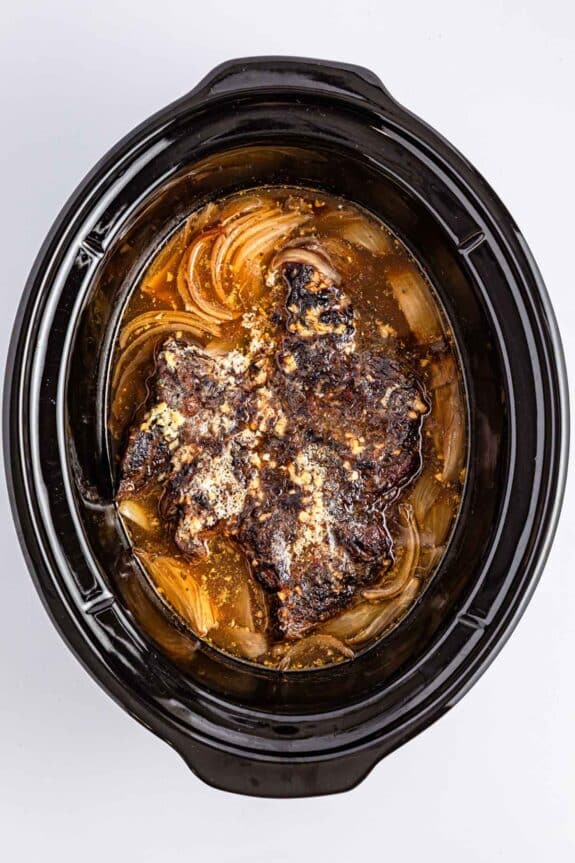 A cooked beef roast with onions and broth inside a black slow cooker.