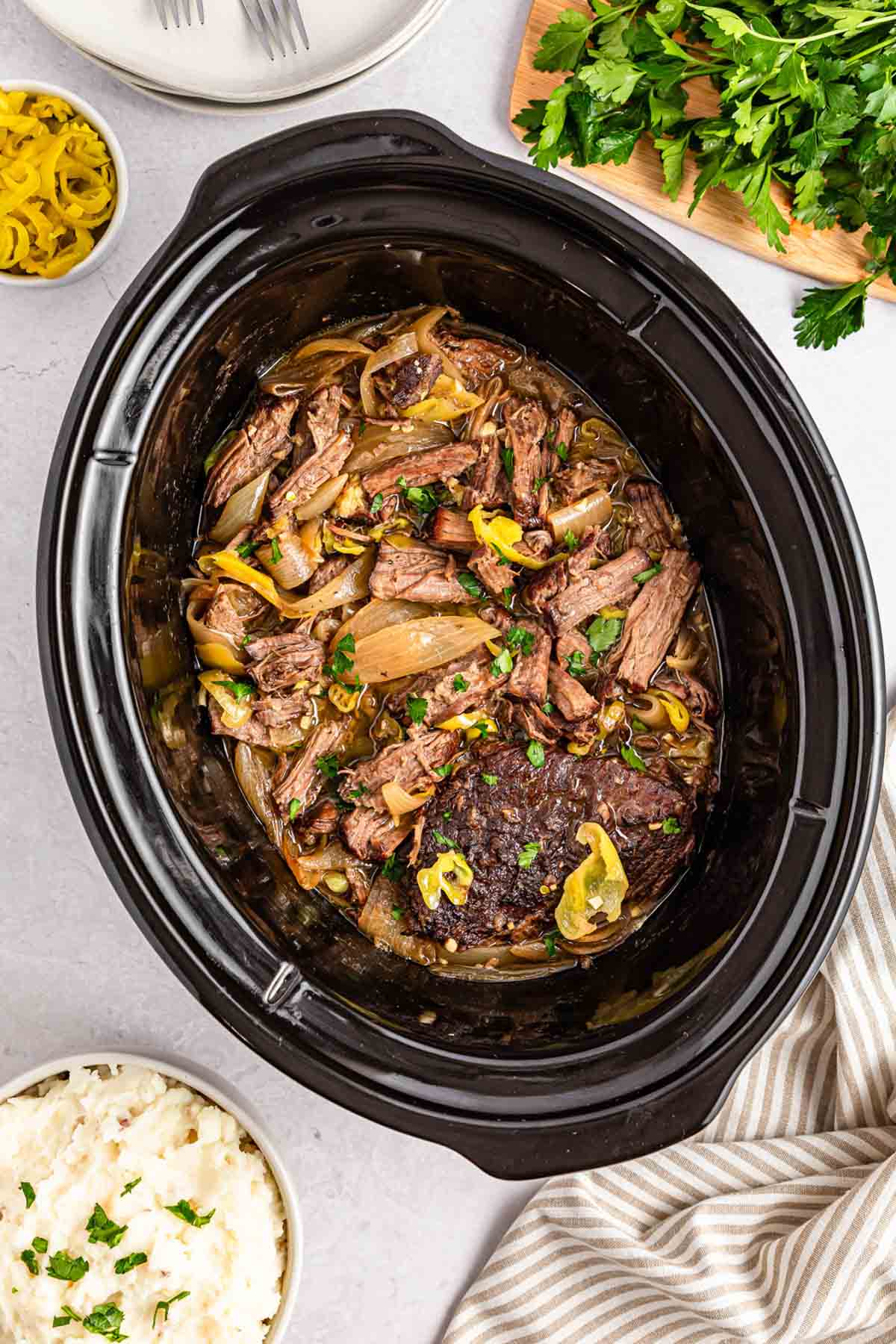 Slow cooker mississippi pot roast with sliced beef, onions, and peppers in a black crock, surrounded by plates, parsley, and mashed potatoes.