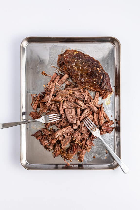 A stainless steel tray holds shredded beef and a whole piece of cooked beef, reminiscent of a classic slow-cooker Mississippi pot roast, accompanied by two forks.