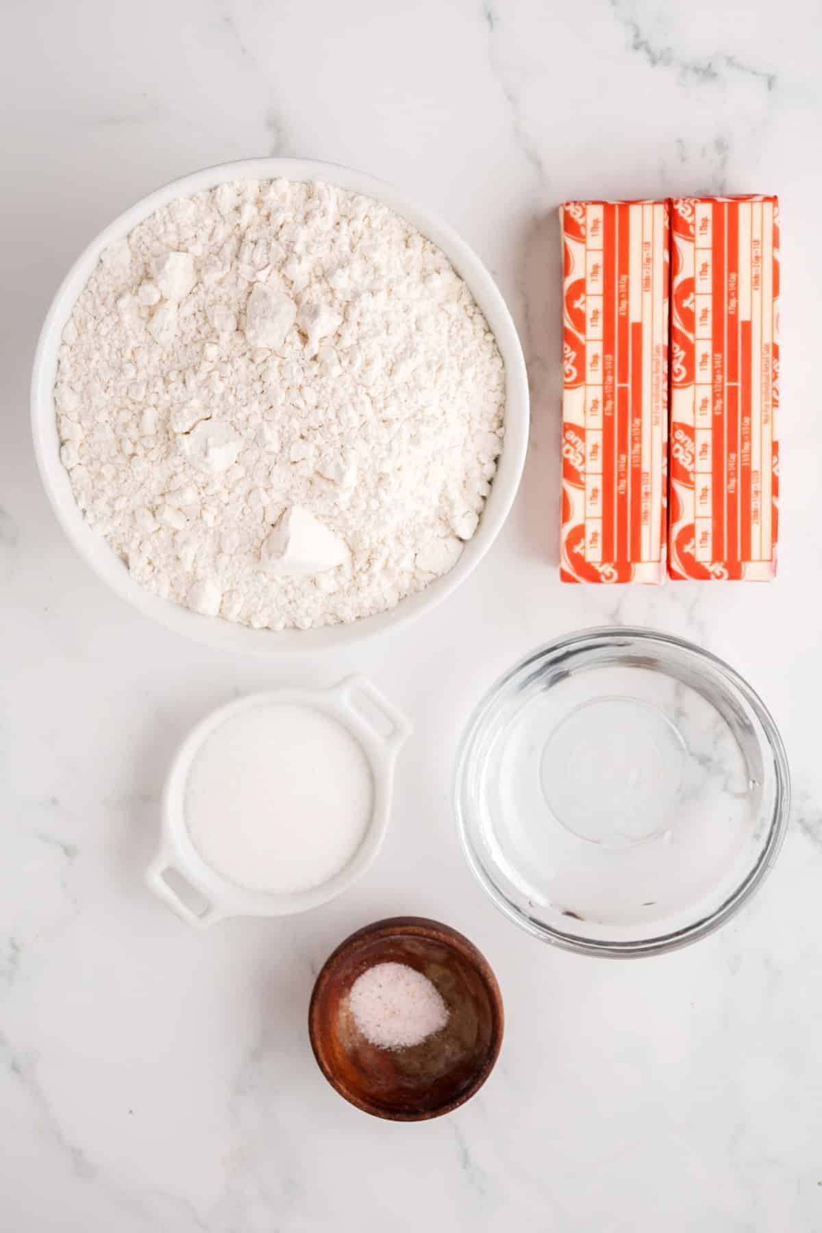 Ingredients for Coconut Pie recipe are the following: a bowl of flour, two sticks of butter, a small bowl of sugar, a small bowl of salt, and a bowl of water arranged on a marble surface.