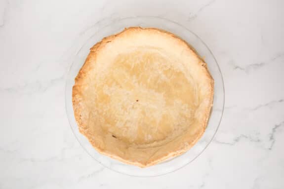 An empty, baked pie crust in a clear glass pie dish on a white marble surface for the Coconut Pie.