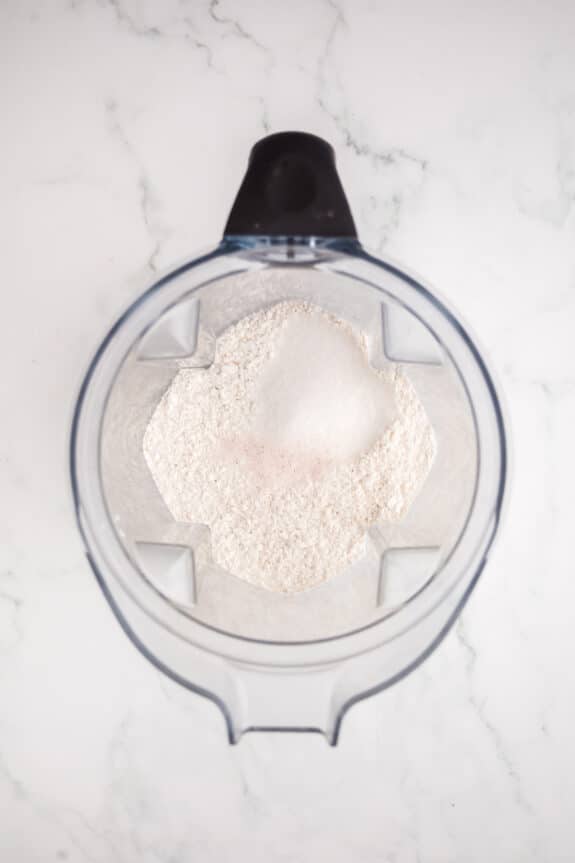 Top view of a blender containing flour, sugar, and a small amount of pink salt on a white marble surface for Coconut Pie recipe