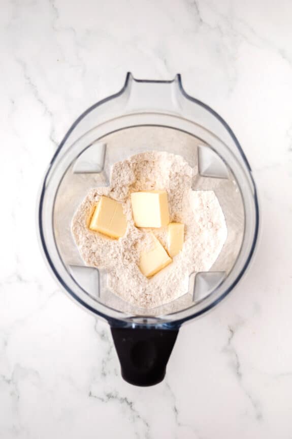 Overhead view of a blender containing flour and several cubes of butter on a white marble surface for the Coconut Pie recipe