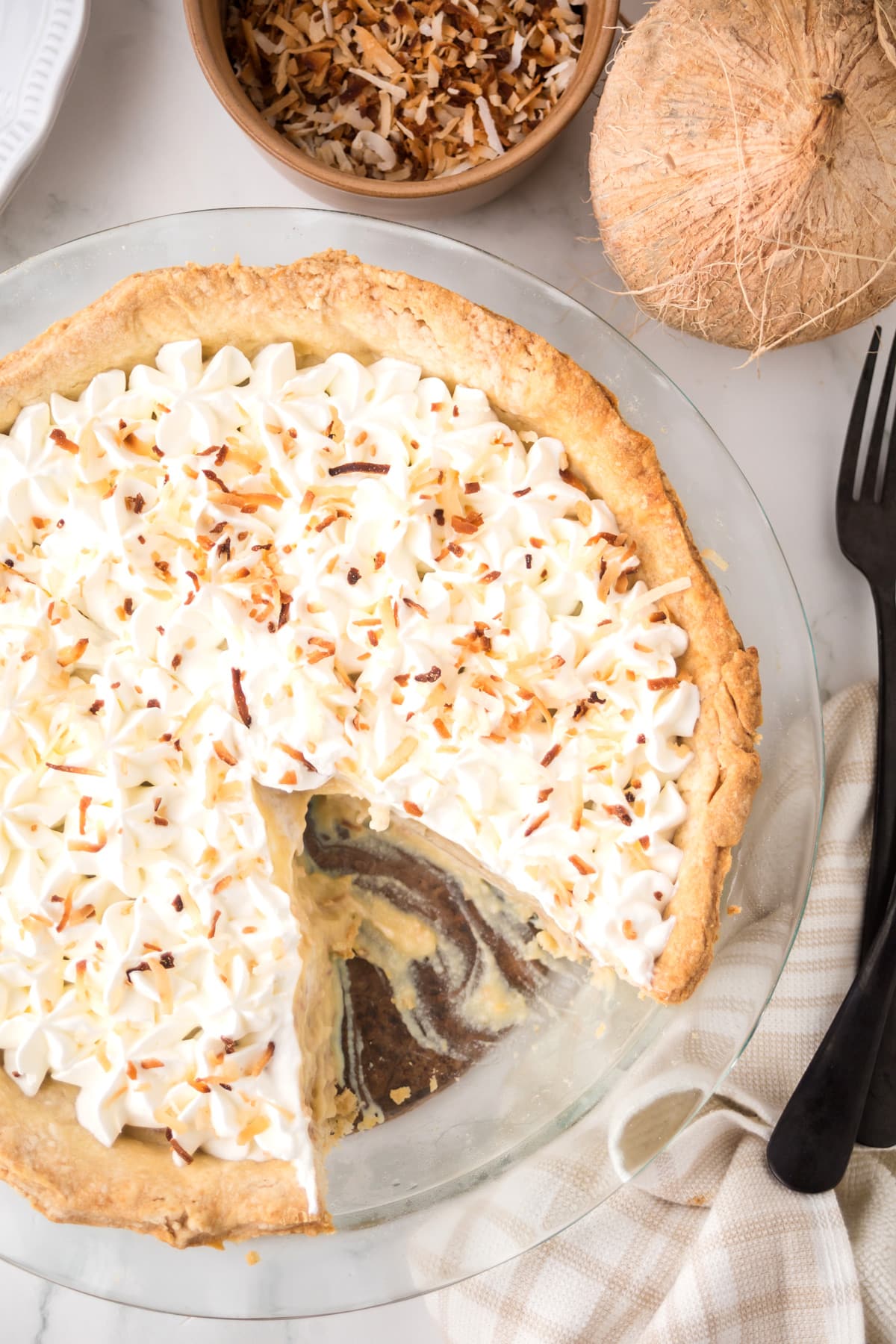 A coconut cream pie with a slice missing, topped with whipped cream and toasted coconut, sits on a glass pie plate next to a bowl of toasted coconut and a whole coconut.