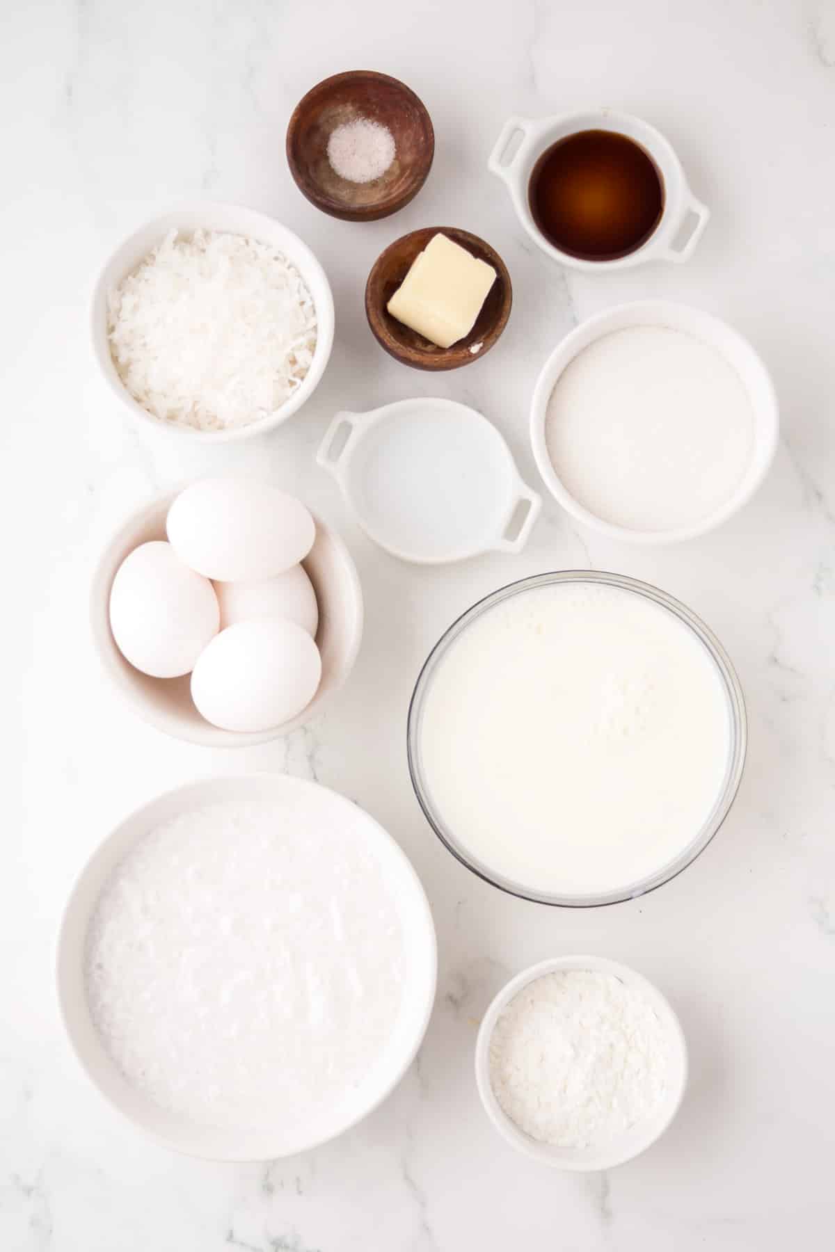 Various baking ingredients including eggs, milk, sugar, shredded coconut, flour, salt, butter, vanilla extract, and water arranged in bowls on a white surface.
