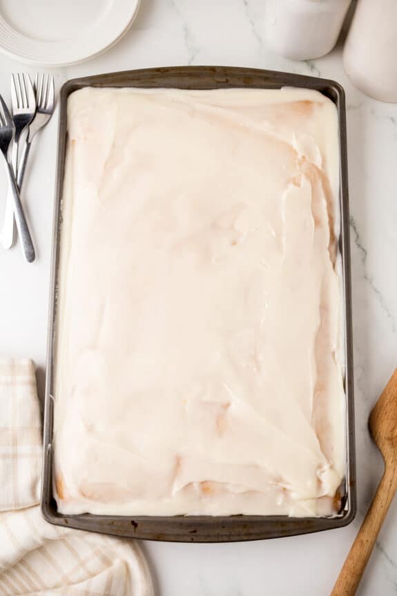 A large rectangular White Texas Sheet Cake covered in white icing on a metal baking pan, with forks, plates, a towel, and a wooden spatula nearby.