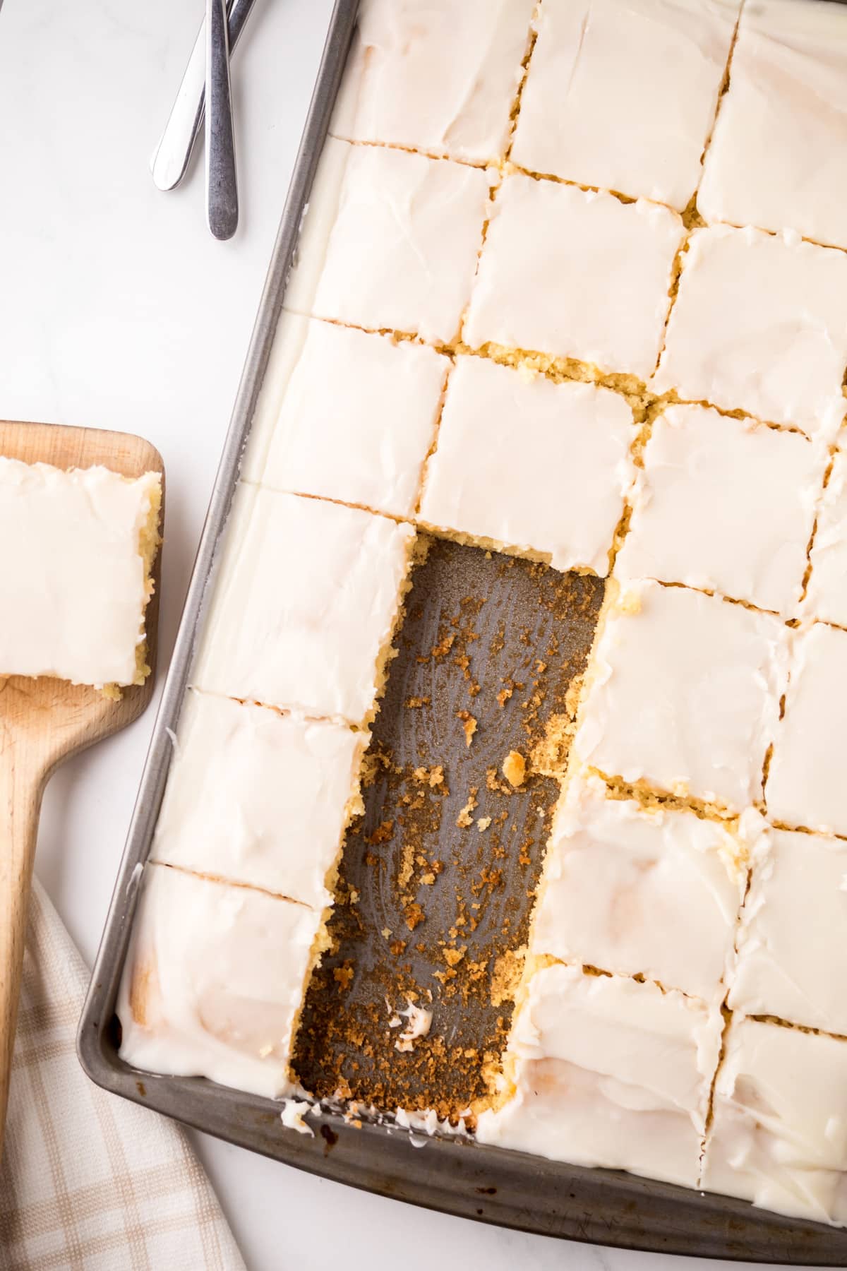 A baking sheet with evenly cut frosted White Texas Sheet Cake, one piece missing, and a single bar on a wooden spatula next to the pan.