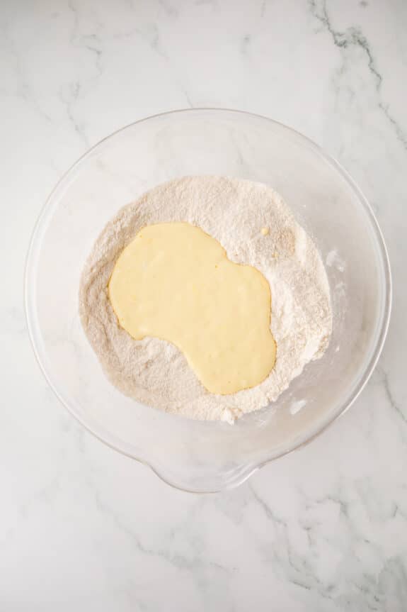 A clear mixing bowl containing eggs, sour cream, and almond extract for White Texas Sheet Cake recipe on a marble counter.