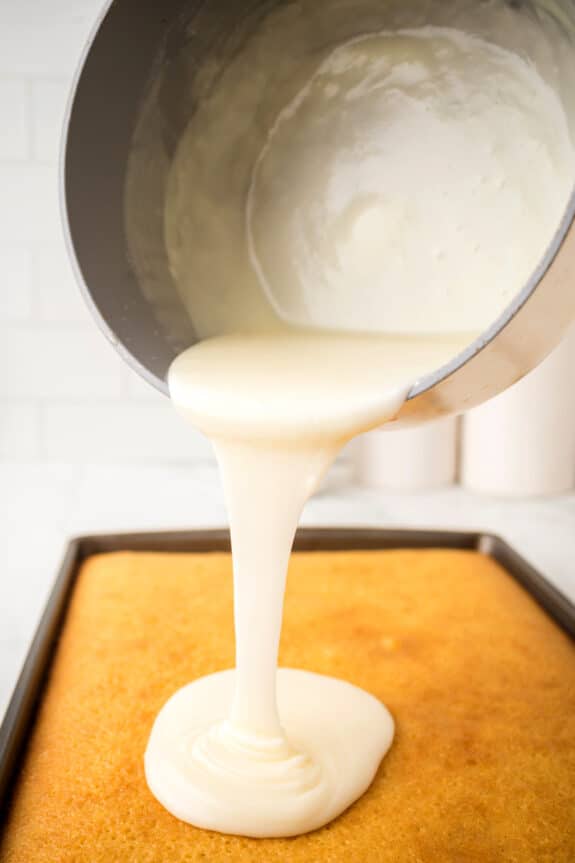 A pot of white icing is being poured onto a rectangular sheet cake. This is one process in preparing White Texas Sheet Cake