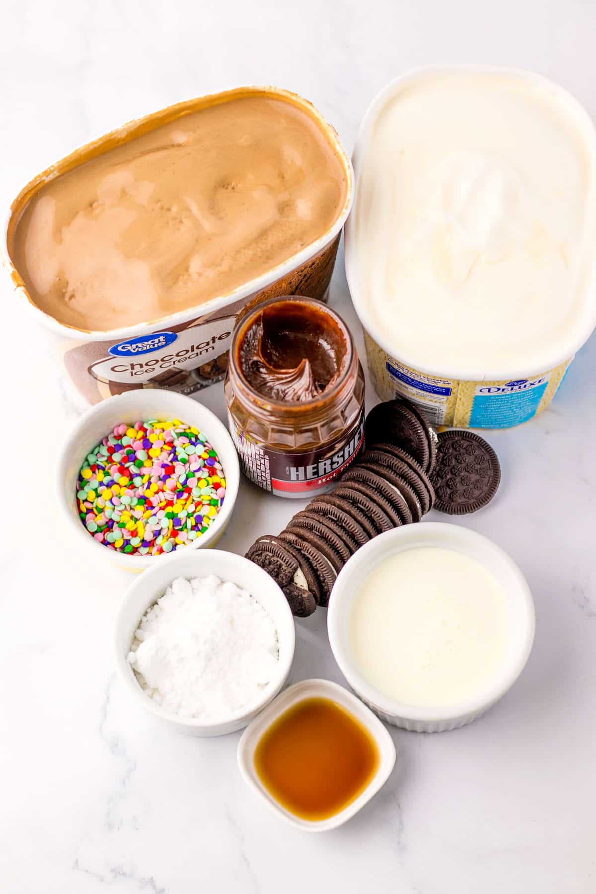 Top-down view of ingredients for Dairy Queen Ice Cream Cake includes: ice cream, chocolate cream cookies, jarred hot fudge, heavy whipping cream, powdered sugar, vanilla extract, and sprinkles.