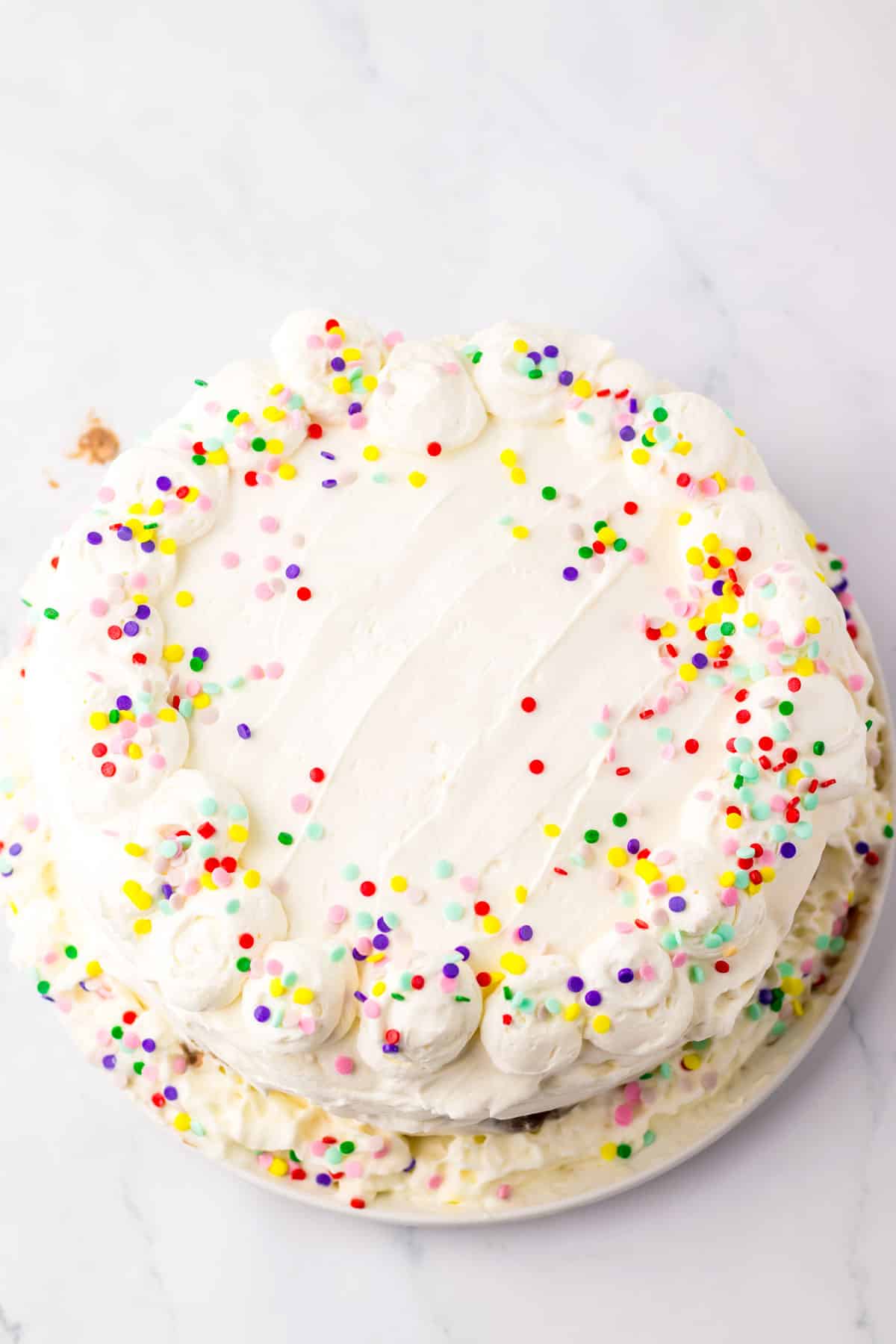 Dairy Queen Ice Cream Cake with white frosting and colorful sprinkles, decorated with piped frosting swirls around the edge, on a white plate.