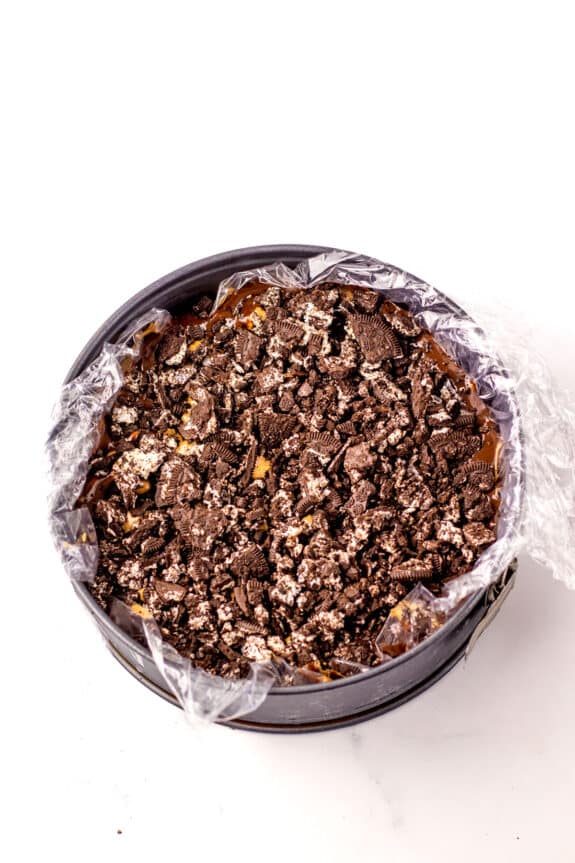 A round baking pan lined with plastic wrap, filled with a dessert topped with crushed oreos for Dairy Queen Ice Cream Cake.
