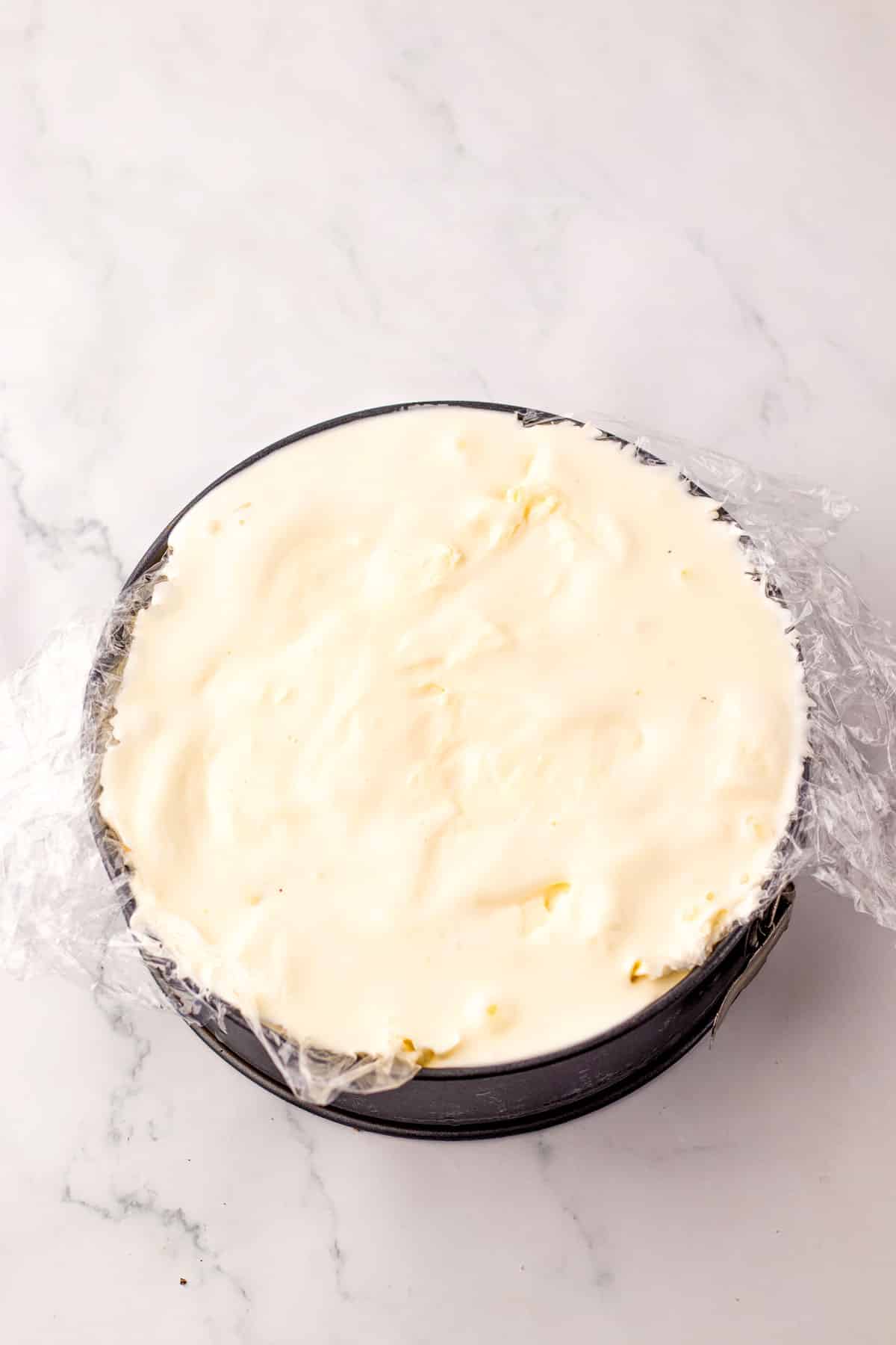 Dairy Queen Ice Cream Cake filled with a smooth, creamy white frosting, partially covered with plastic wrap, sits on a marble countertop.