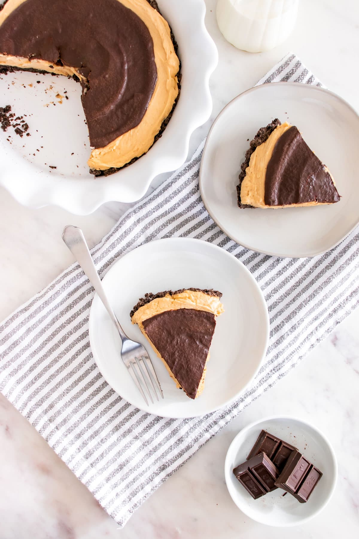 Overhead view of two plates, each with a slice of chocolate-topped, Buckeye Pie with a cookie crust, next to a pie dish and a small bowl of chocolate pieces.