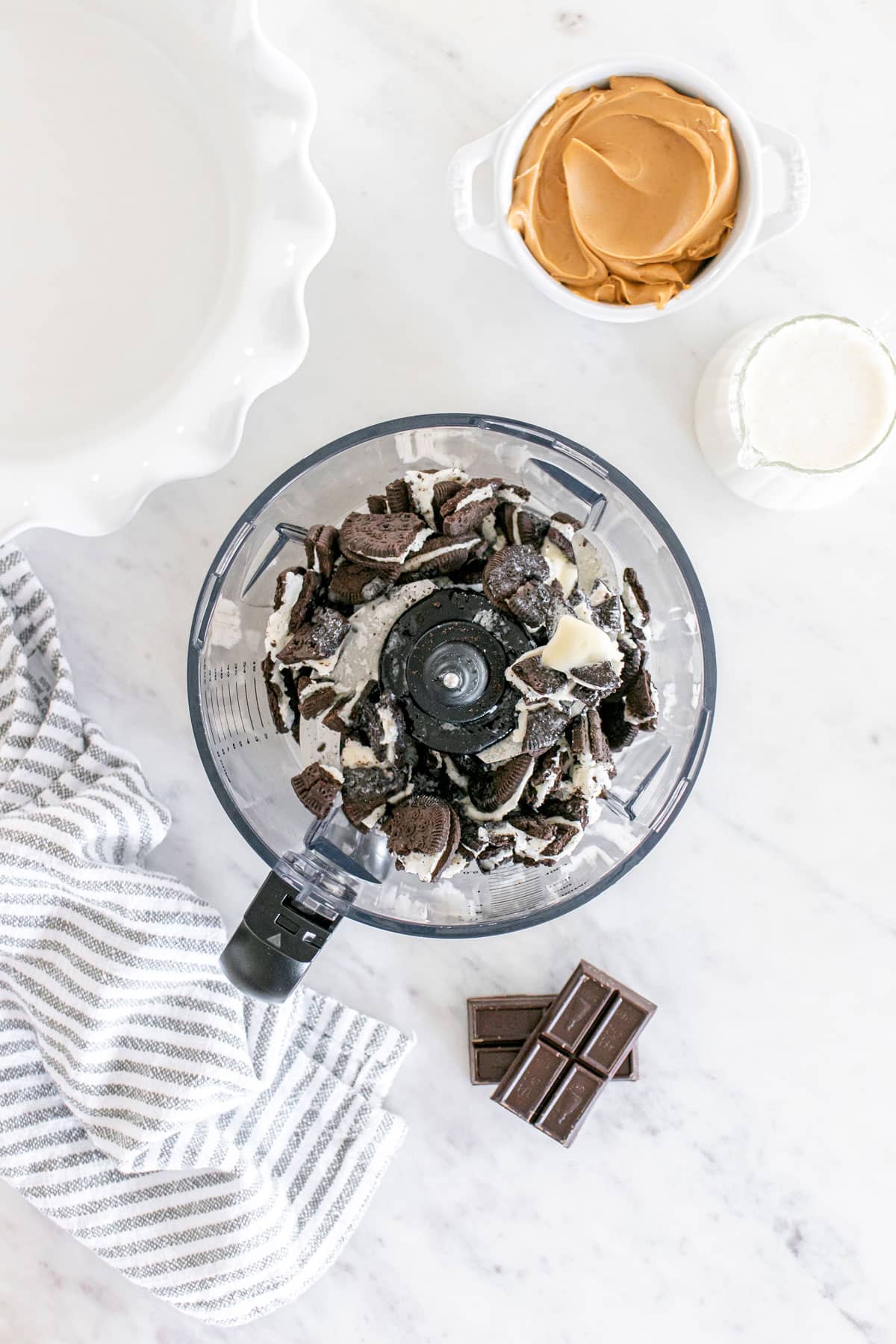 A food processor with crushed oreo cookies, a bowl of peanut butter, a cup of sugar, chocolate pieces, and a striped towel on a marble countertop- a process in preparing Buckeye Pie