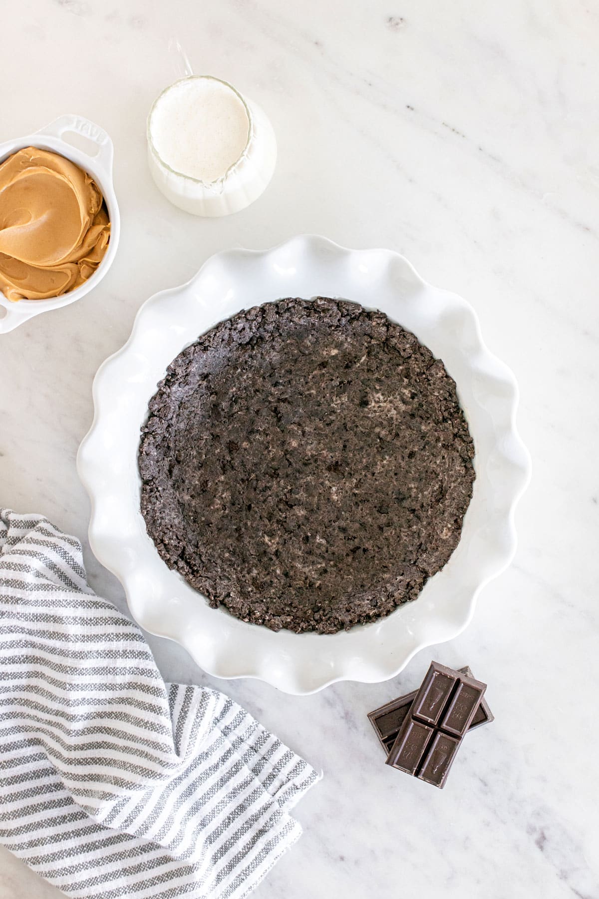 A chocolate cookie crust in a white pie dish on a marble surface, with a bowl of peanut butter, a cup of milk, a chocolate bar, and a striped cloth nearby for Buckeye Pie recipe