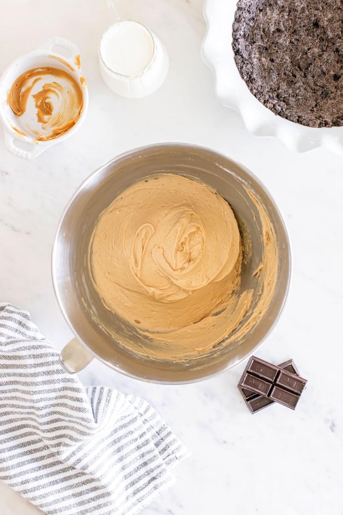 A metal mixing bowl with creamy batter, a chocolate bar, a striped kitchen towel, an empty dish with residue, a milk pitcher, and a pie pan with a dark crust on a marble surface for Buckeye Pie recipe.