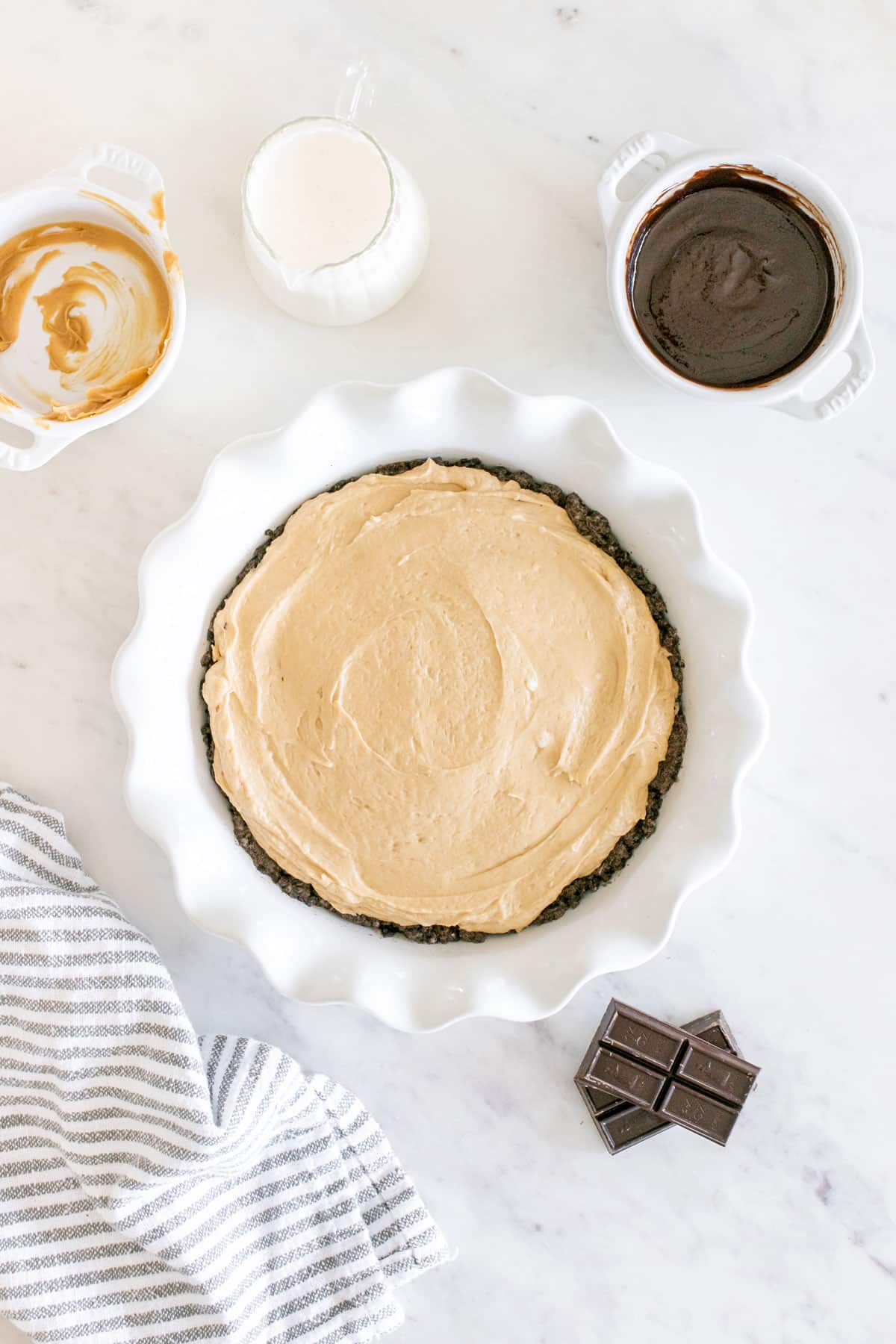 Buckeye Pie with a creamy filling in a white dish, surrounded by bowls of caramel, chocolate sauce, a jug of cream, a striped cloth, and pieces of chocolate on a marble surface.
