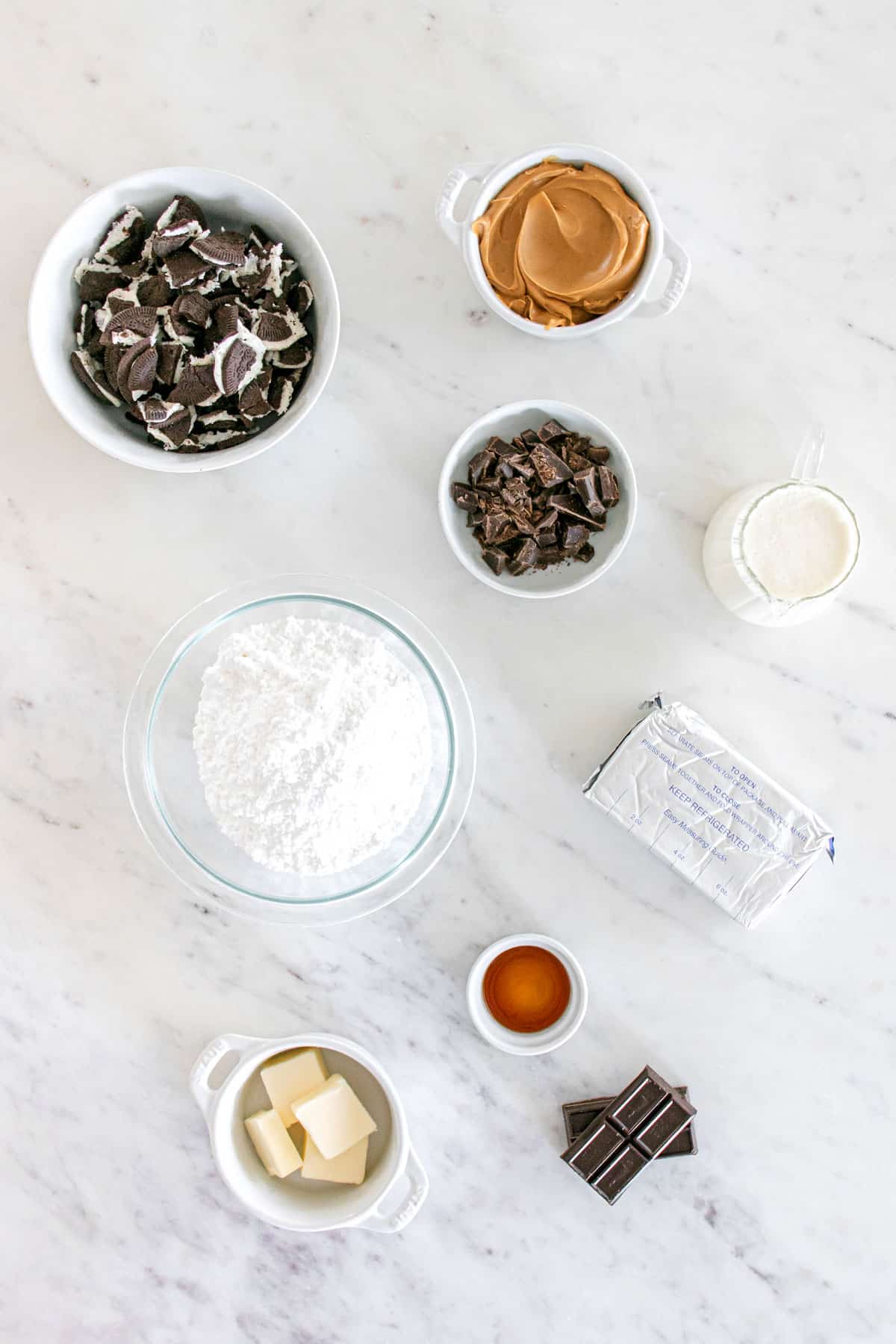 Assorted baking ingredients for Buckeye Pie on a marble surface, including crushed oreo cookies, peanut butter, cream cheese, chocolate bar, powdered sugar, cream cheese, butter, whipping cream, and vanilla extract in bowls and containers.