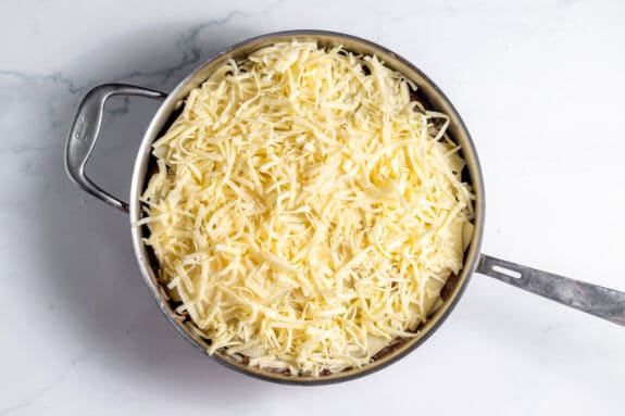 A metal pan filled with shredded cheese on top of an unbaked casserole, placed on a white marble surface.